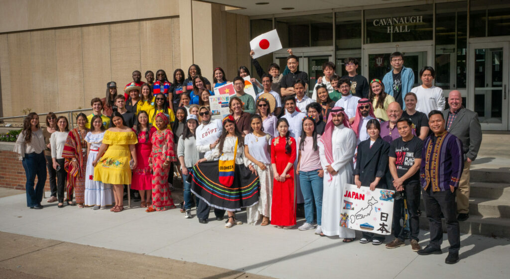 Picture of a group of people standing on the steps of a building wearing various clothing that represents their country and are from many different countries.