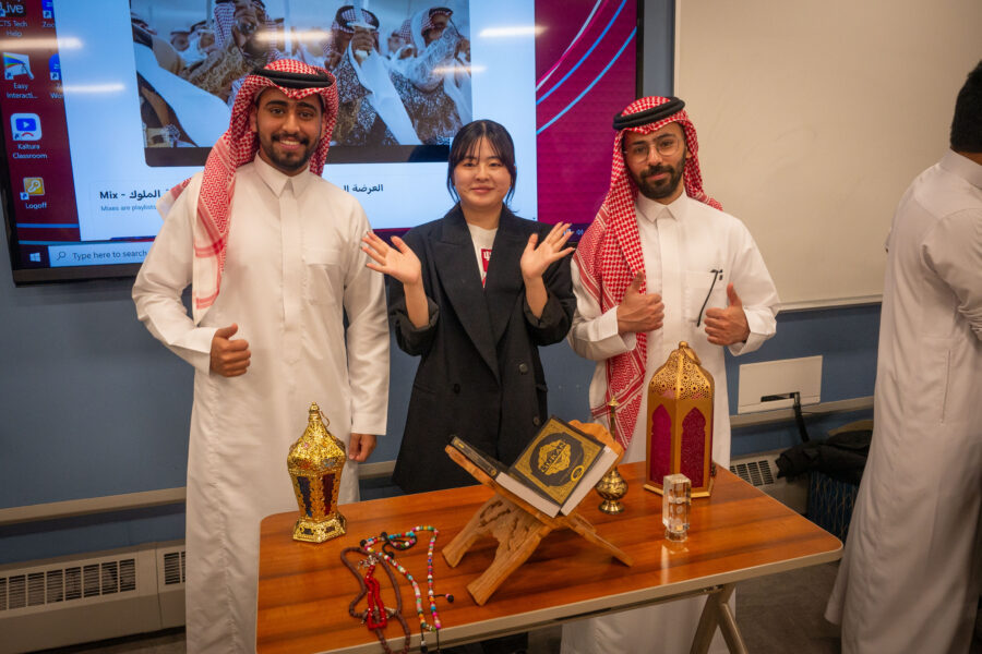 Picture of three individuals, two men dressed in traditional clothing from their country with a woman formally dressed standing between them standing behind a table with objects from their country in front of them.