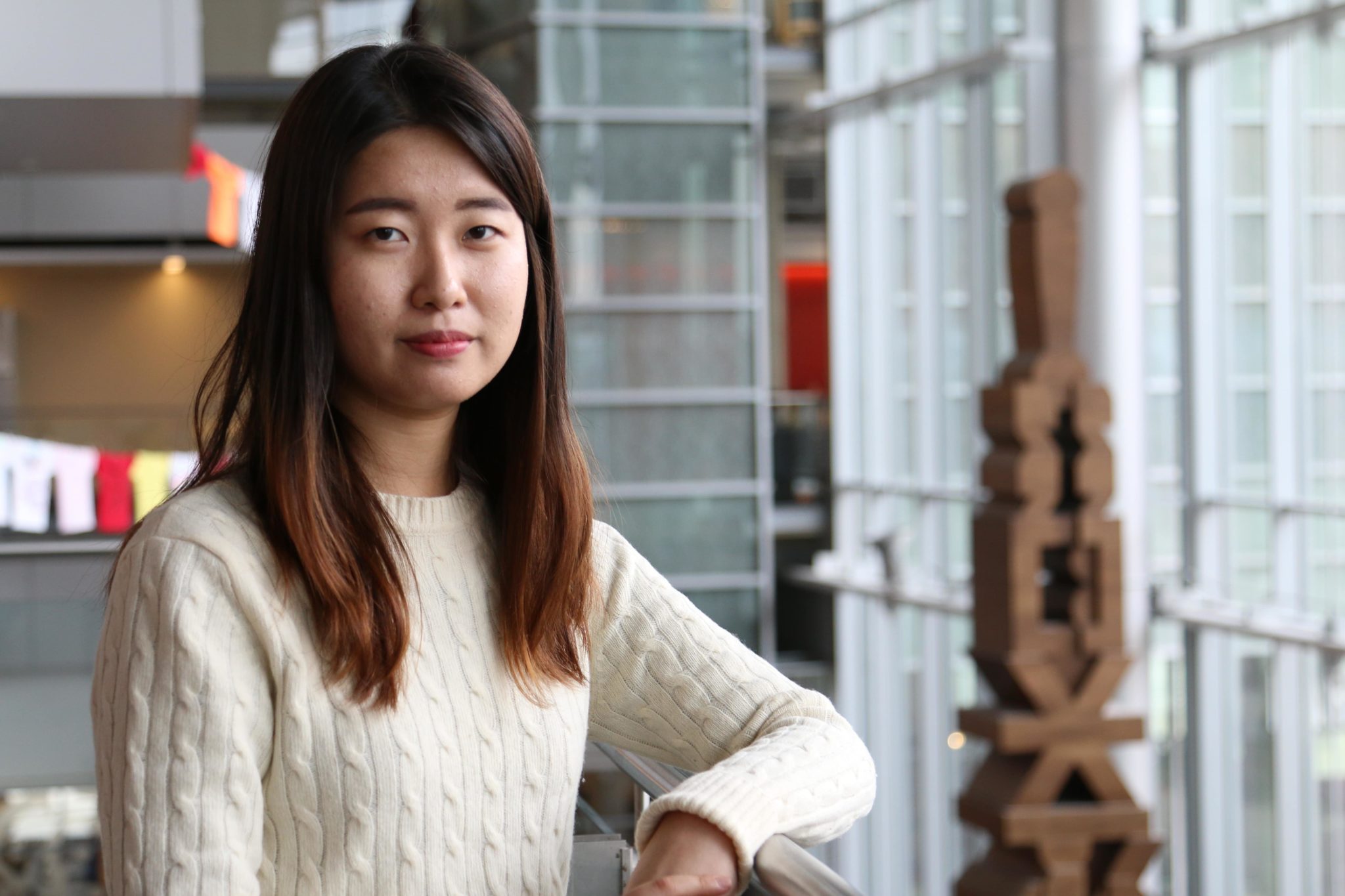 young woman in a cream sweater standing indoors by a railing in a modern building with large windows and contemporary decor